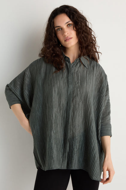 A woman with curly dark hair wears the Pleated Easy Shirt, an oversized, textured blouse in dark gray, styled with black pants against a plain light background.