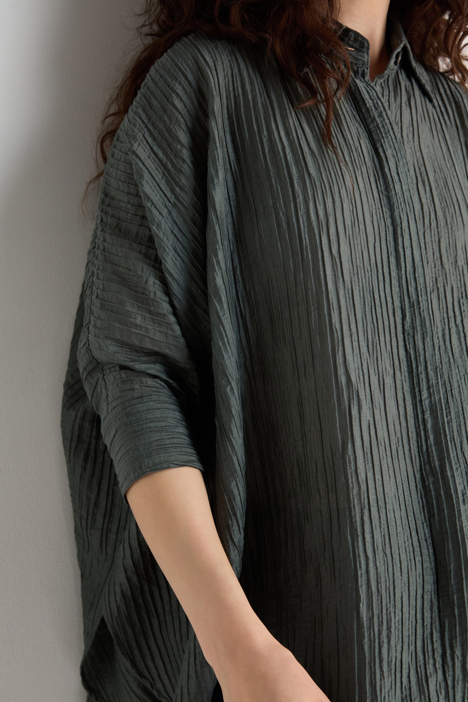 A person wearing the Pleated Easy Shirt in dark gray, featuring three-quarter sleeves and textured pleats, stands against a light wall; only the torso and part of the arm are visible.