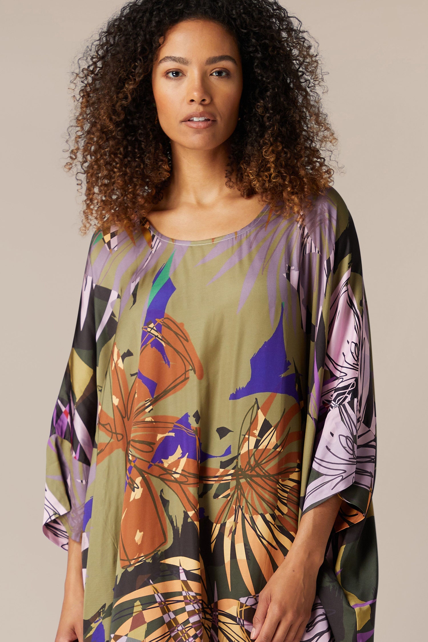 A woman with curly hair wearing a vibrant Jungle Kaftan stares directly at the camera, standing against a plain beige background.