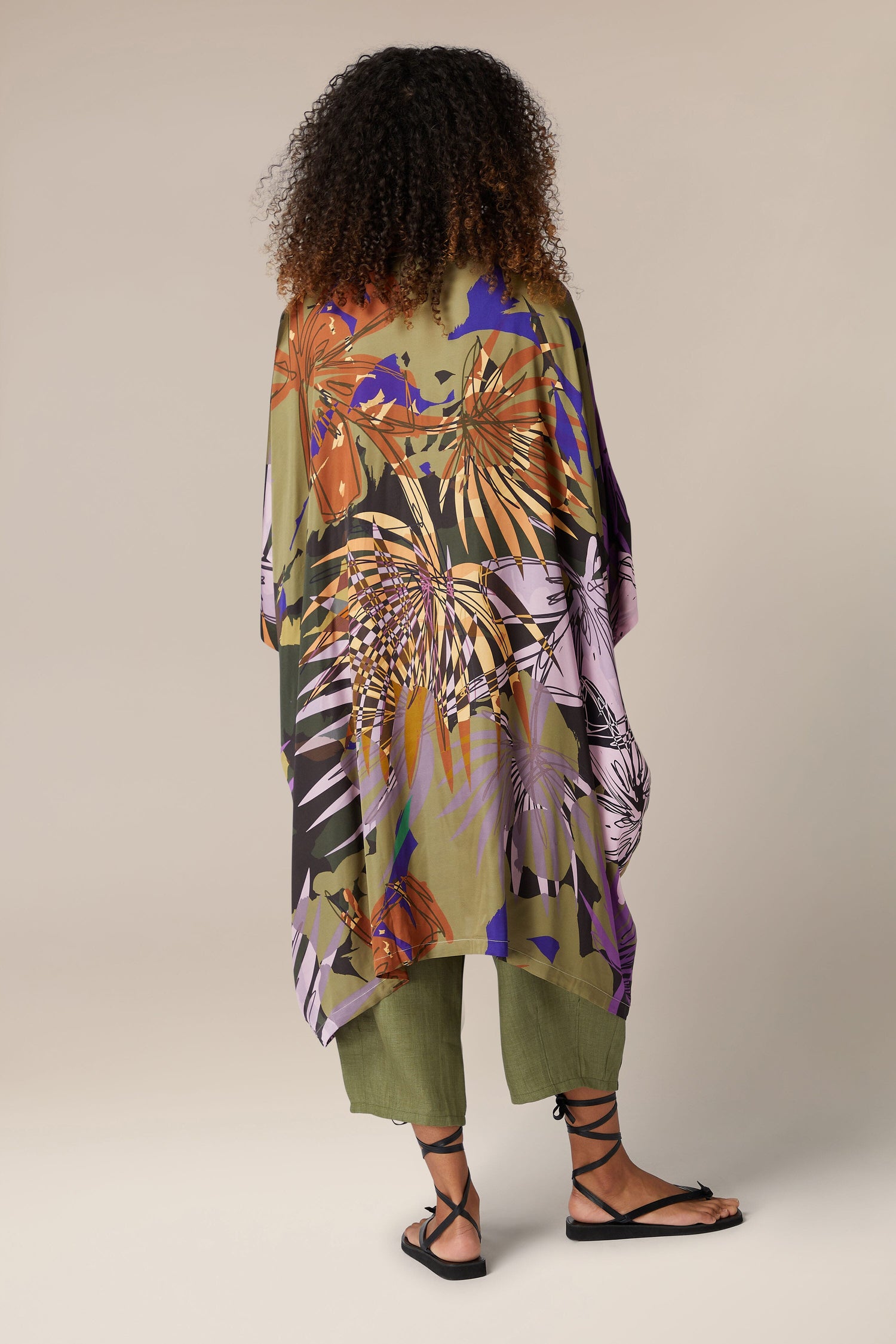 A person stands facing away, showcasing a colorful floral-patterned Jungle Kaftan with elegant draping over green pants and black sandals against a plain background.