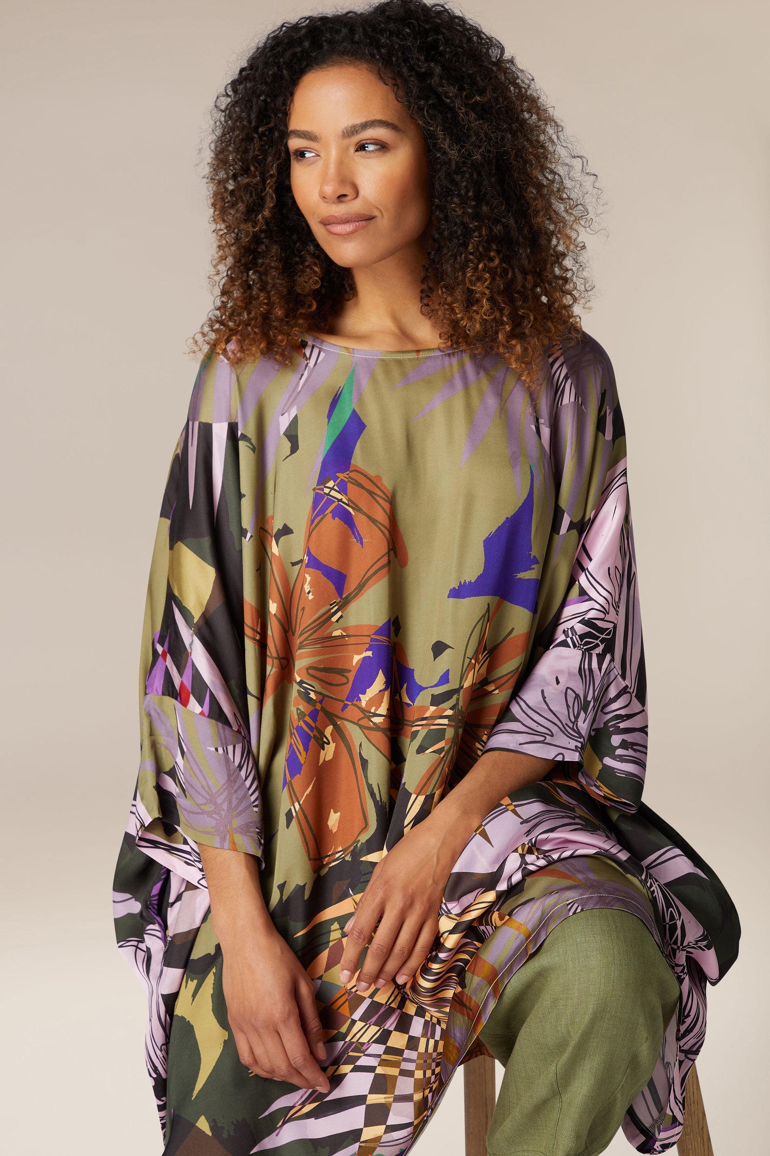 A woman with curly hair wearing a vibrant Jungle Kaftan and green pants is seated and looking to her right against a plain background.