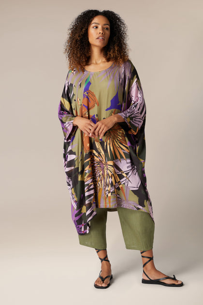 A person with curly hair wears a Jungle Kaftan over green pants and black sandals, standing against a neutral background.