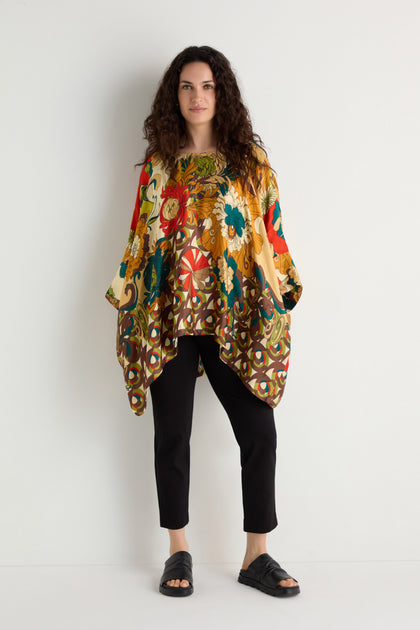 A woman stands against a white wall wearing the Japanese Bloom Tunic featuring a vibrant floral print, paired with black pants and sandals.