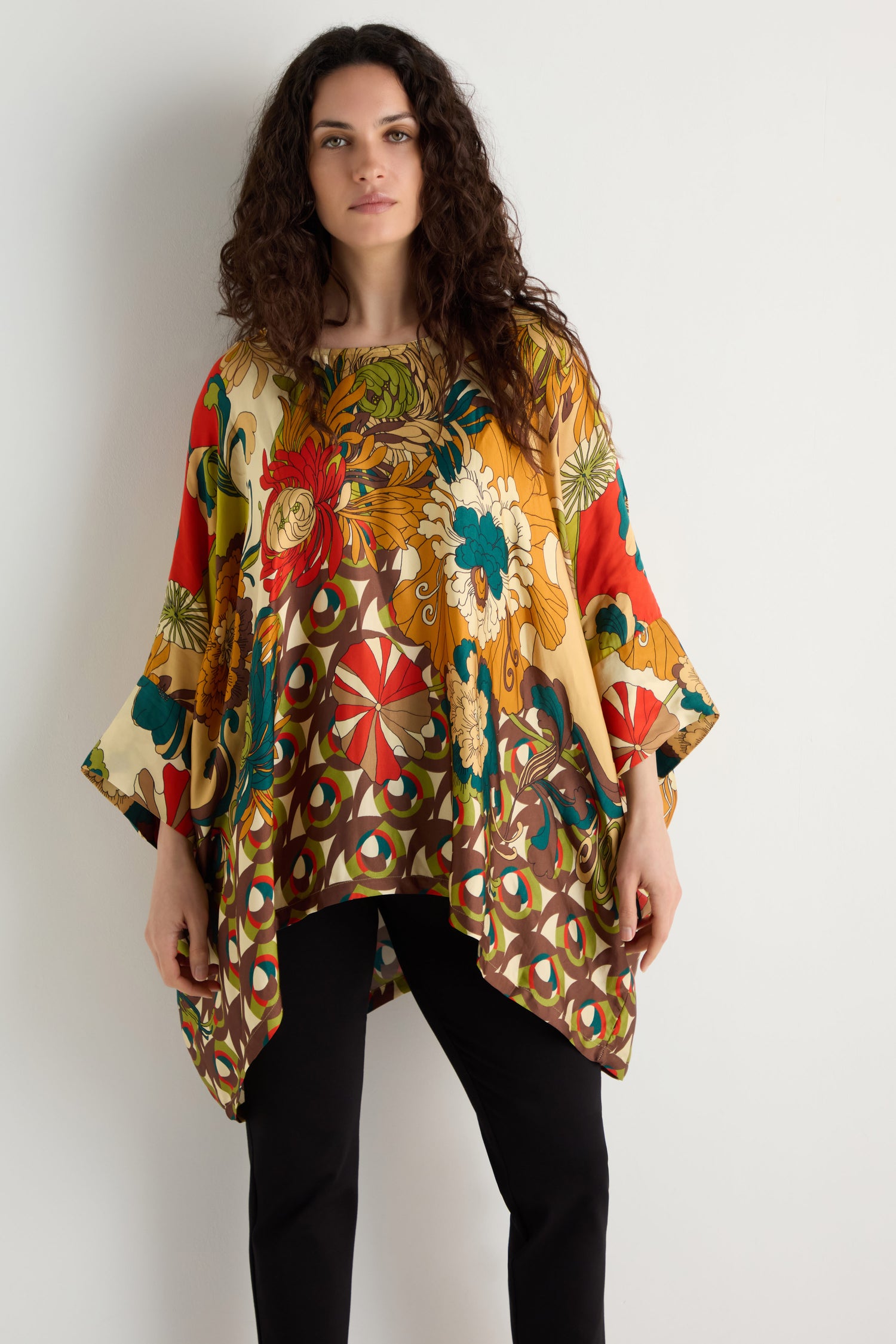 Woman stands against a white wall wearing the Japanese Bloom Tunic, featuring a vibrant floral print and wide sleeves, paired with black pants as she looks directly at the camera.