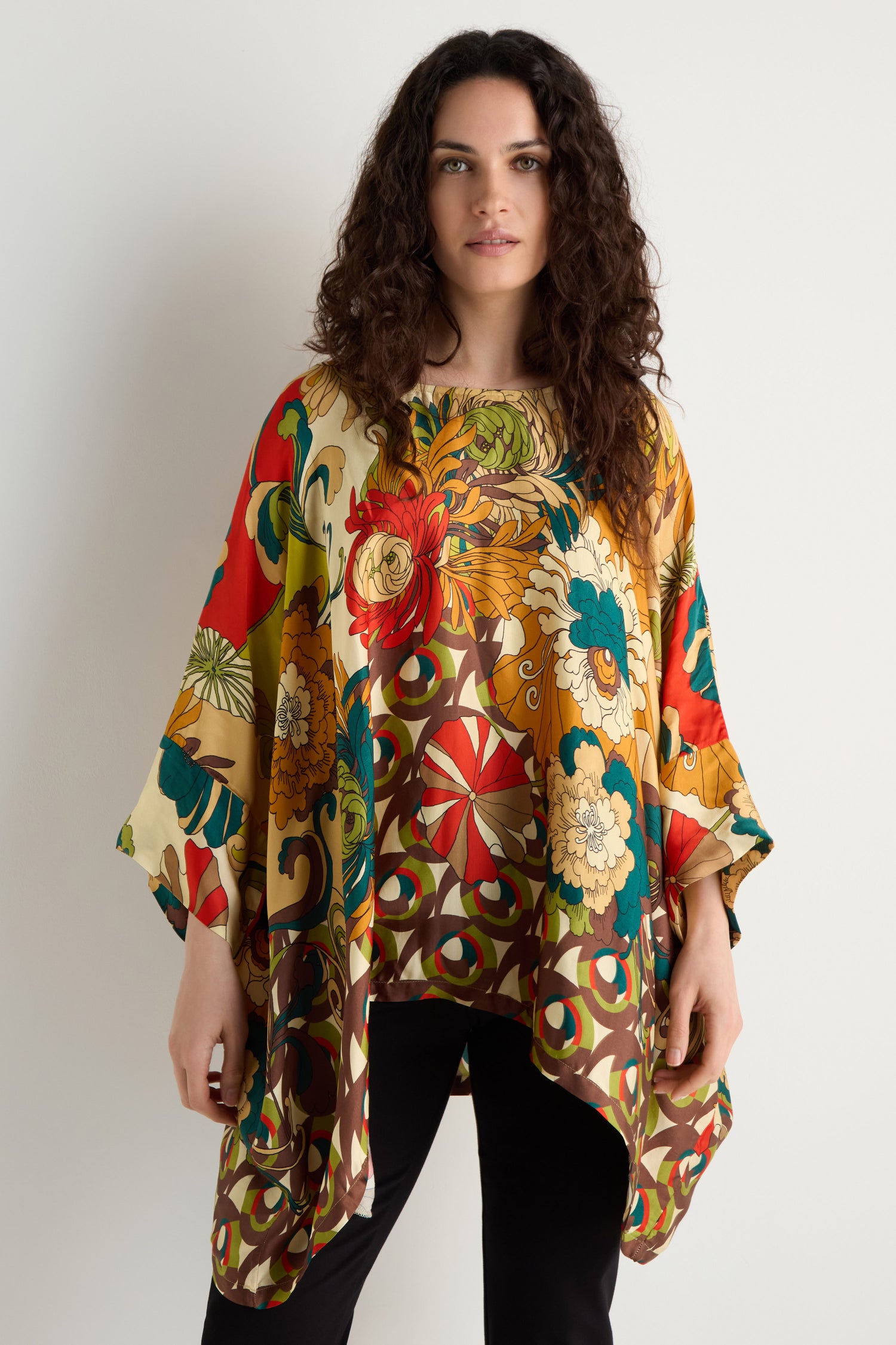 A woman stands against a white wall, wearing our Japanese Bloom Tunic in a vibrant floral print with wide sleeves, paired with black pants for a relaxed and elegant look.