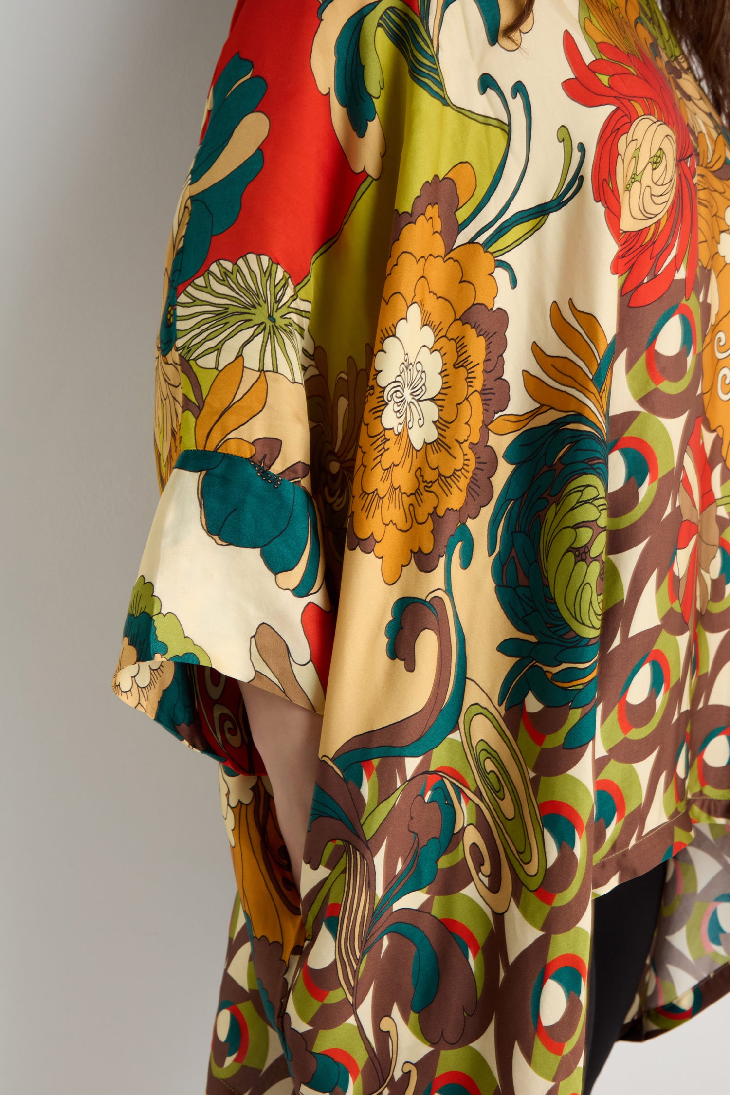 A person wears the Japanese Bloom Tunic, an oversized top with a colorful floral print in red, yellow, green, and brown tones; only the upper body is visible.
