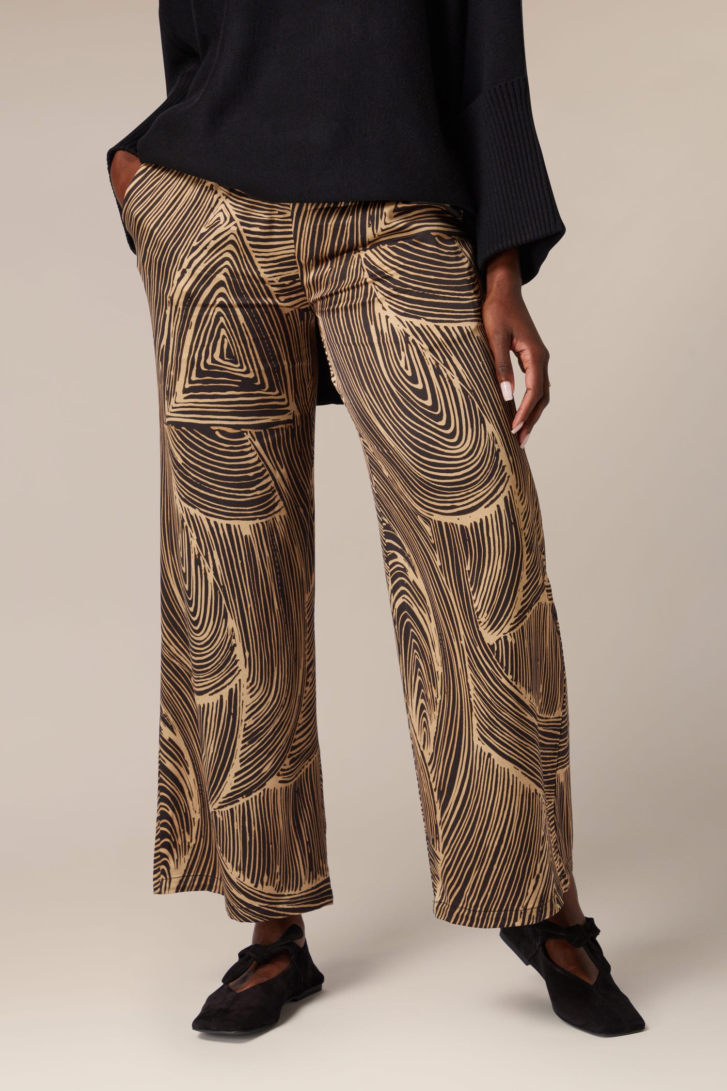 A woman showcasing artisanal flair in her contemporary design Swirl Trouser ensemble, featuring a black top and brown wide leg pants.