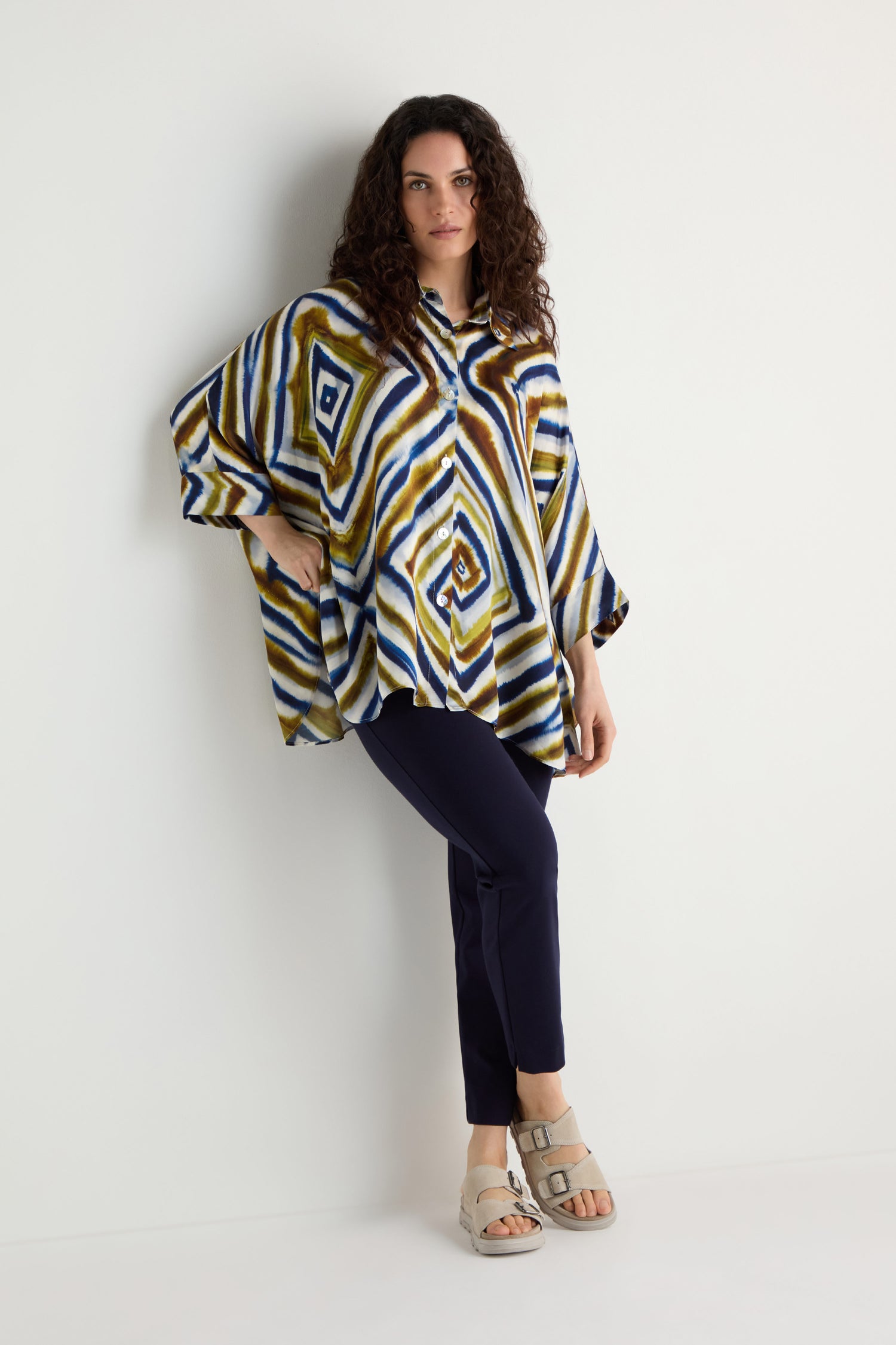 A woman stands against a white wall wearing the Wavy Lines Print Shirt, styled in a loose fit with dark pants and light sandals, one hand on her hip and the other by her side.