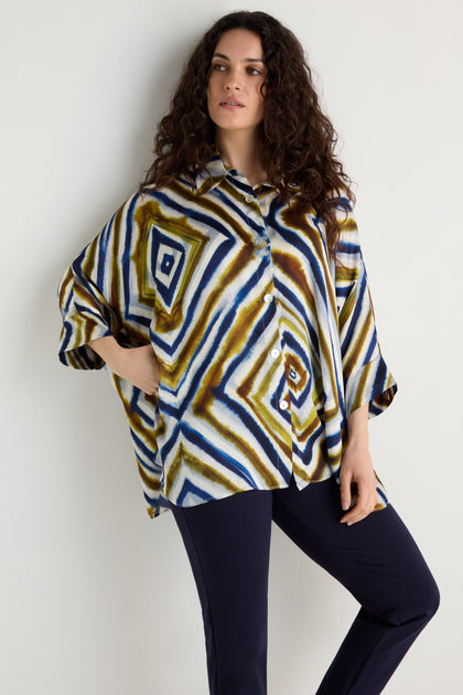 A woman with curly hair wears the Wavy Lines Print Shirt, a loose viscose top with blue, brown, and white abstract wavy patterns, paired with dark pants against a plain background.