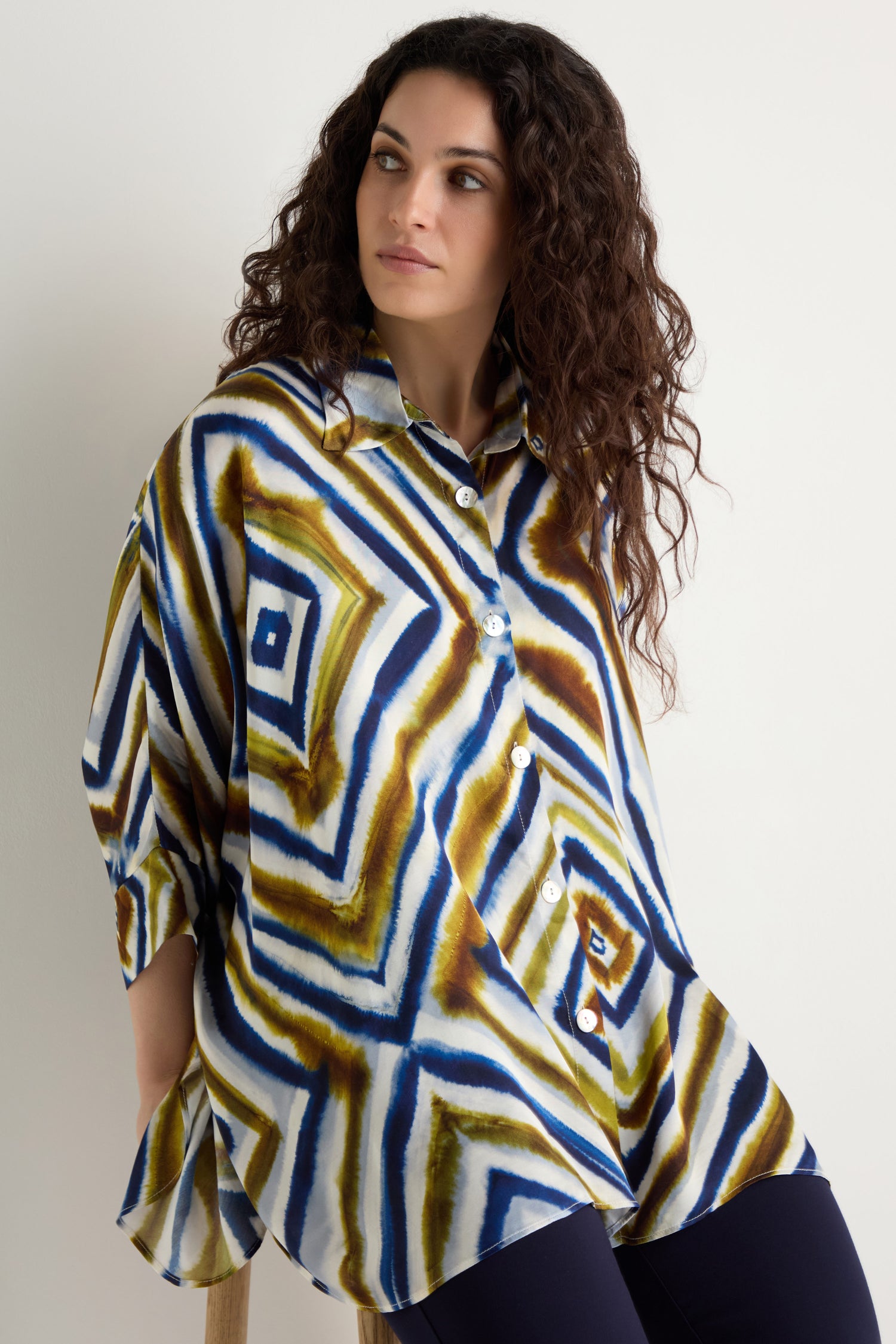 A woman with curly hair sits on a stool wearing the Wavy Lines Print Shirt, featuring geometric blue, green, and brown designs, paired with dark pants.