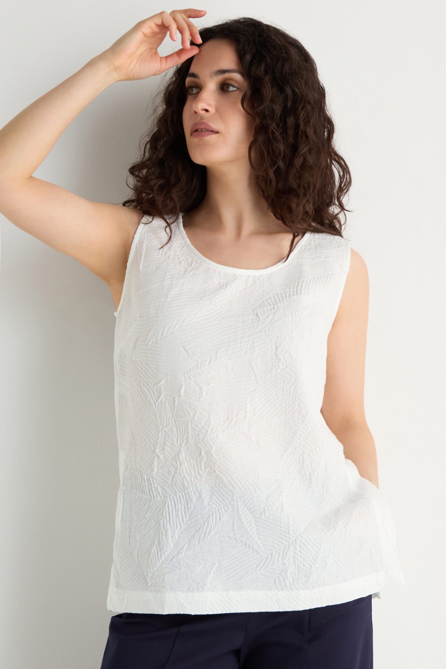 A woman with curly hair models the Pleated Leaf Top, a sleeveless white pleated top, paired with dark pants against a plain light background.