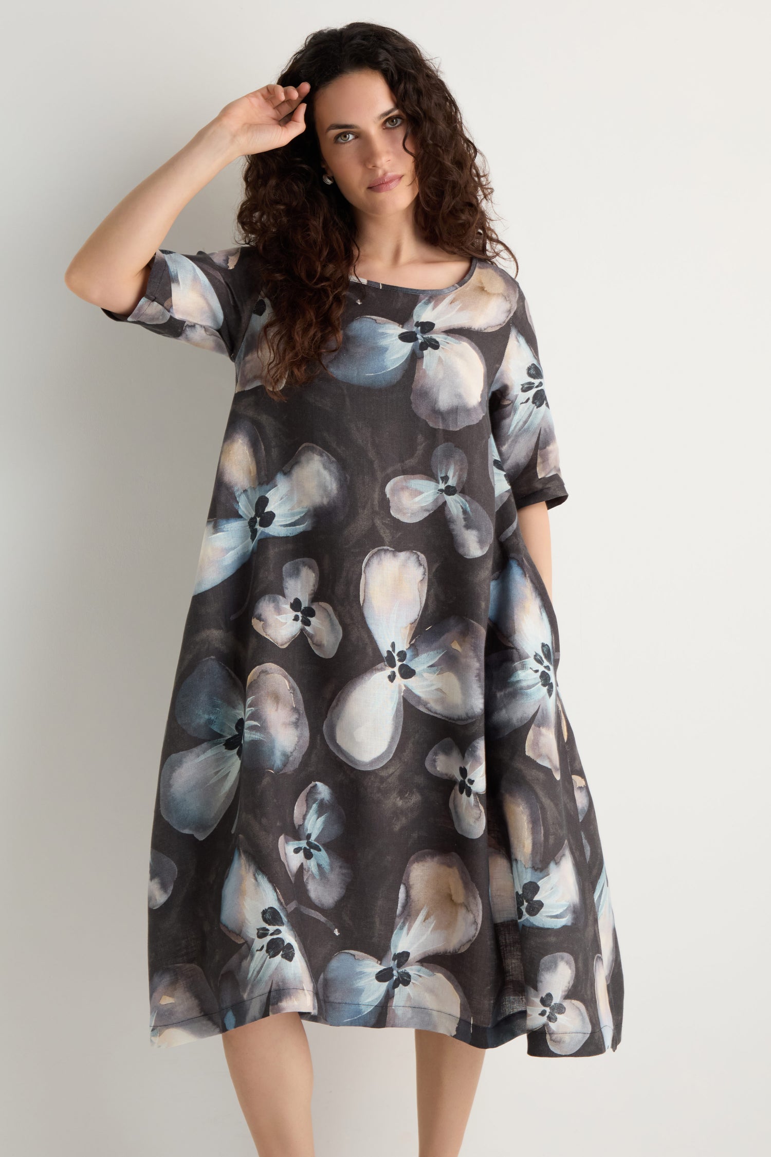 Woman wearing the Abstract Floral Linen Flared Dress, a loose, knee-length piece featuring large abstract floral patterns in blue, white, and beige on a dark base, standing against a plain background.