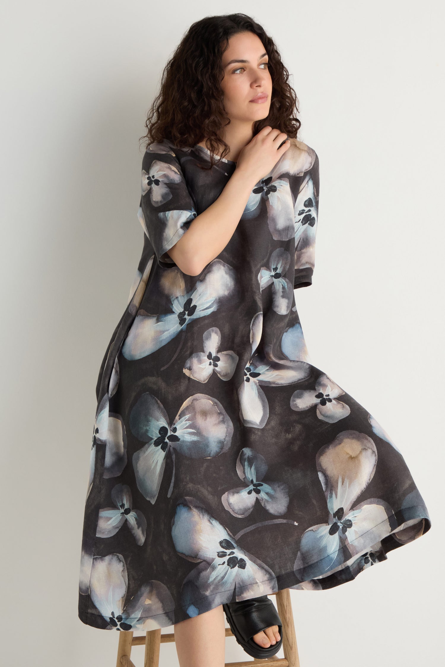 A woman with curly hair sits on a stool against a white background, wearing the Abstract Floral Linen Flared Dress in blue and white with loose, short sleeves and an abstract floral print.