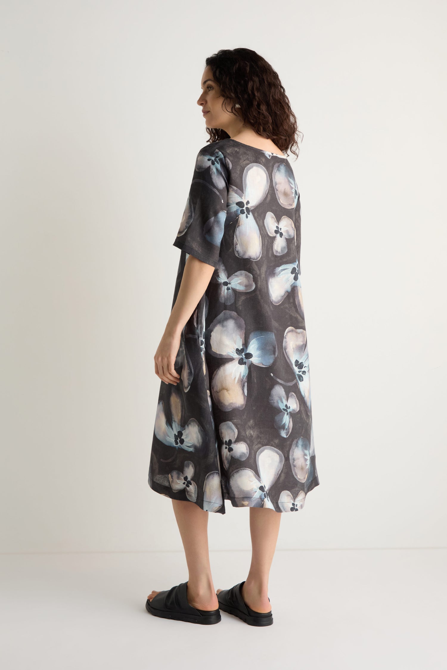 A woman stands sideways in the Abstract Floral Linen Flared Dress, featuring a loose fit and a blue and white abstract floral pattern, paired with black sandals against a plain background.