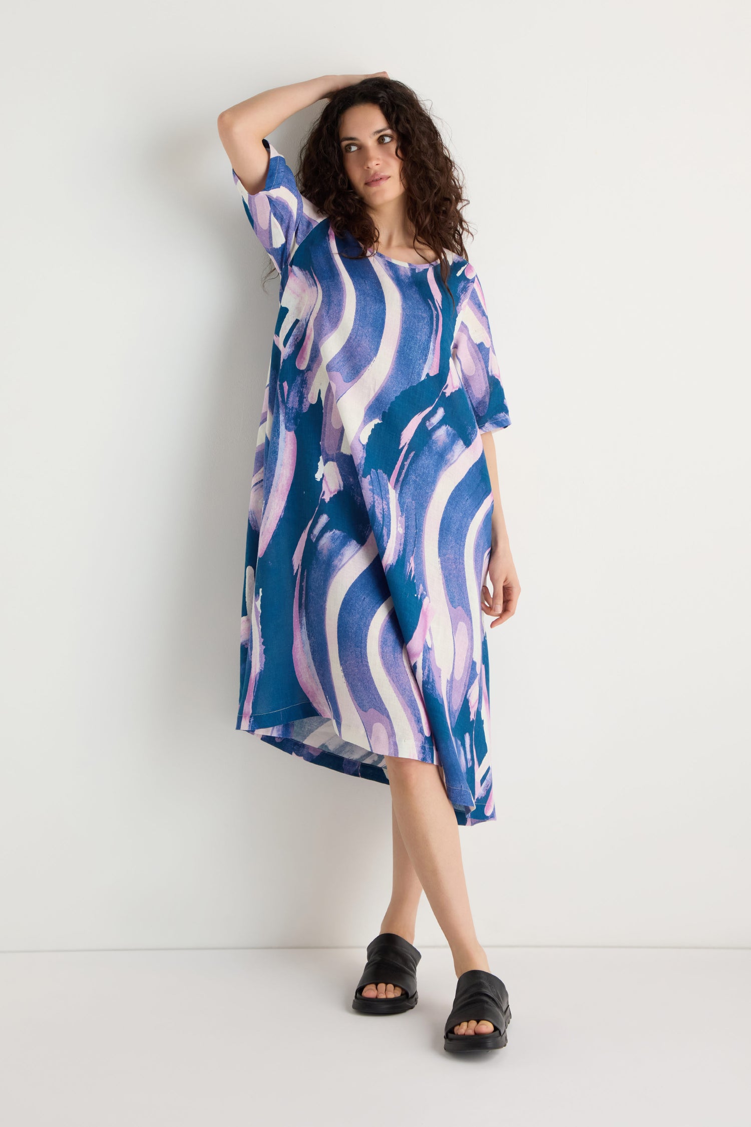 Woman stands against a white wall wearing the Painted Stripes Linen Flared Dress, a loose-fitting midi with blue, purple, and white abstract patterns, paired with black slide sandals.
