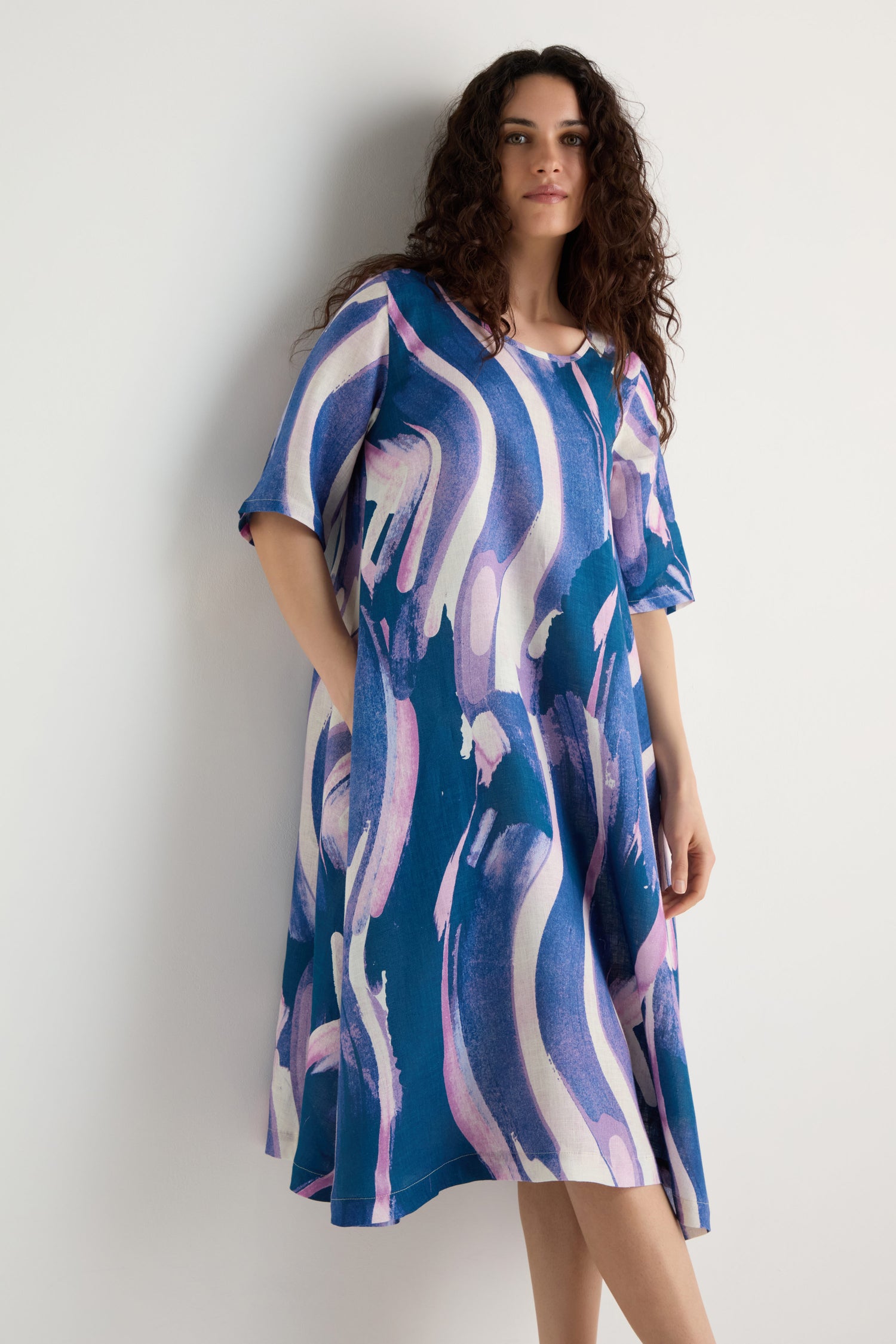 A woman stands against a white wall, wearing the Painted Stripes Linen Flared Dress featuring blue linen with abstract white and purple brushstroke patterns, looking at the camera.