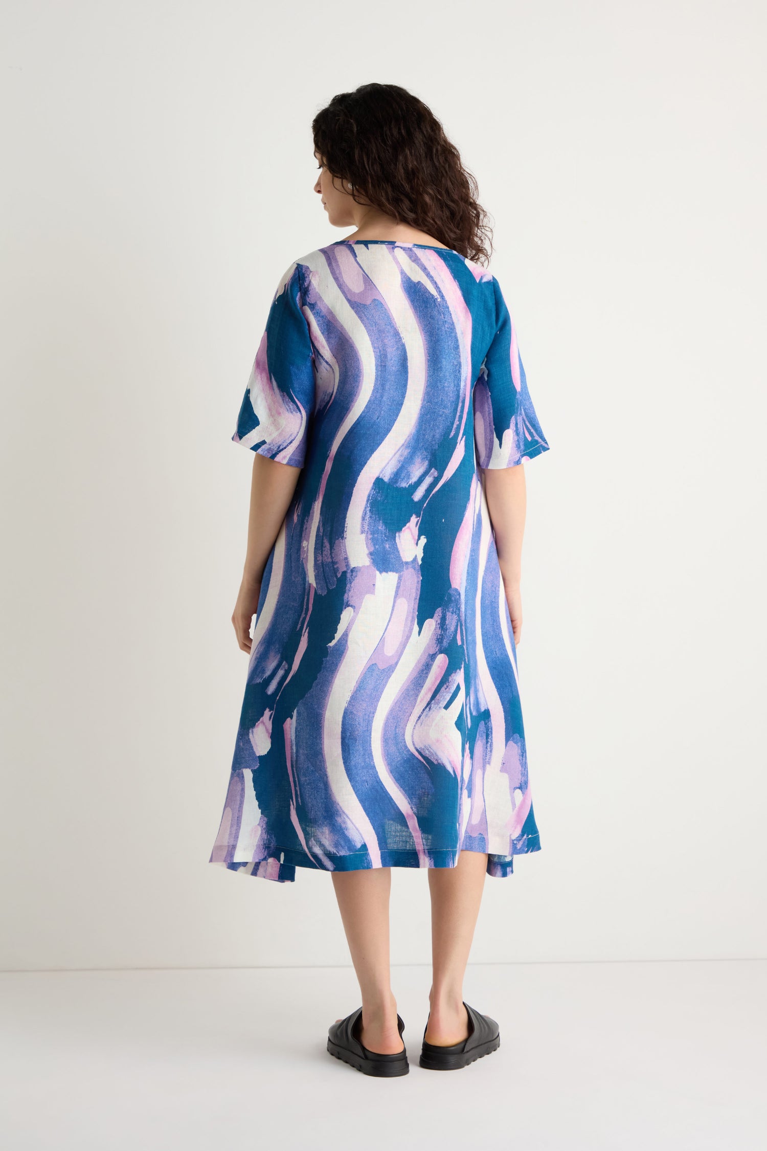 A person stands against a plain background, facing away, wearing the Painted Stripes Linen Flared Dress with blue and purple abstract patterns, paired with black slip-on shoes.