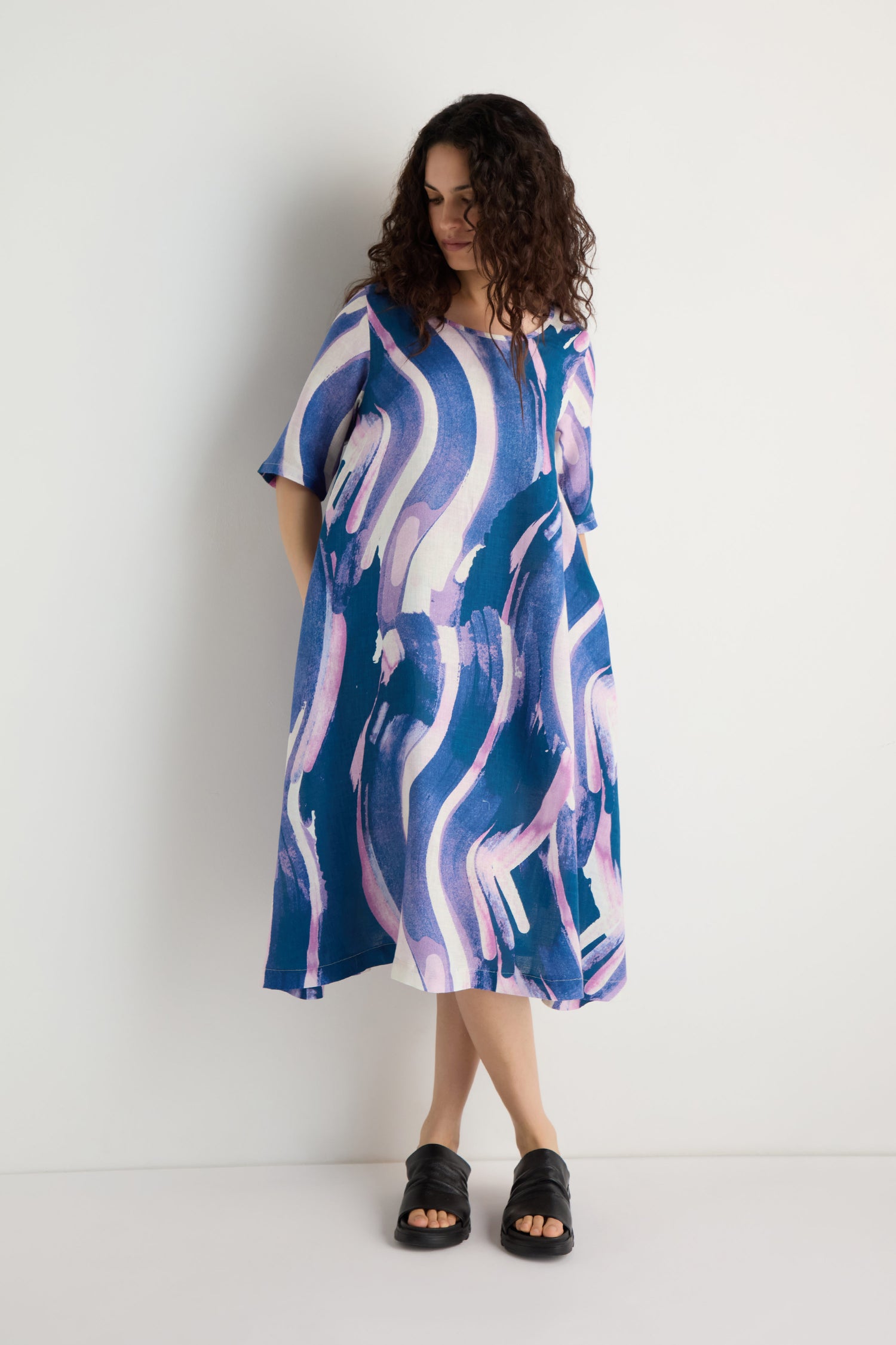 Woman wearing the Painted Stripes Linen Flared Dress, a loose, midi-length piece featuring blue, purple, and white abstract wavy patterns, standing against a plain white background.