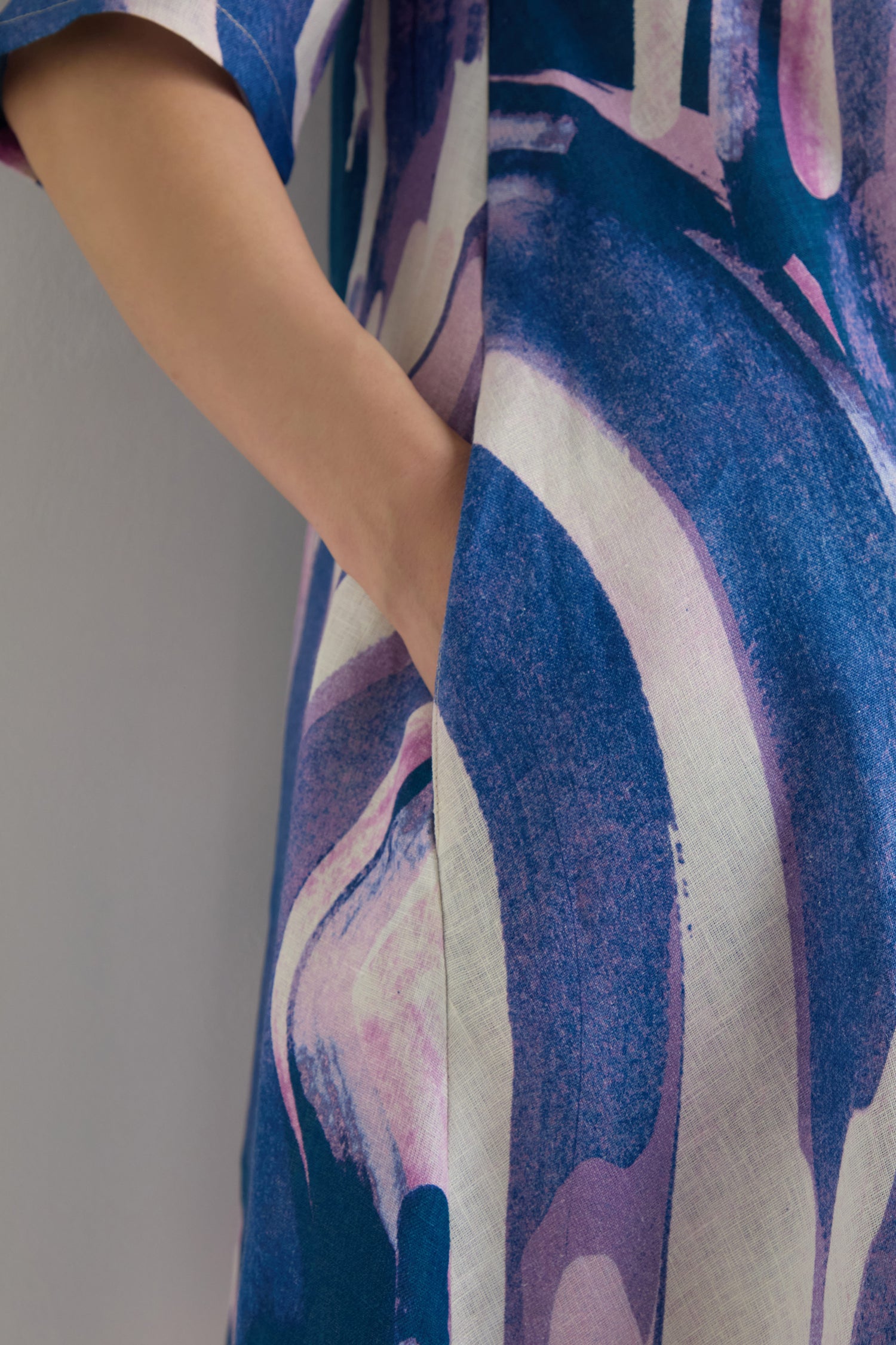 A person stands with their hand in the pocket of the Painted Stripes Linen Flared Dress, a midi-length piece featuring blue and purple painted stripes.