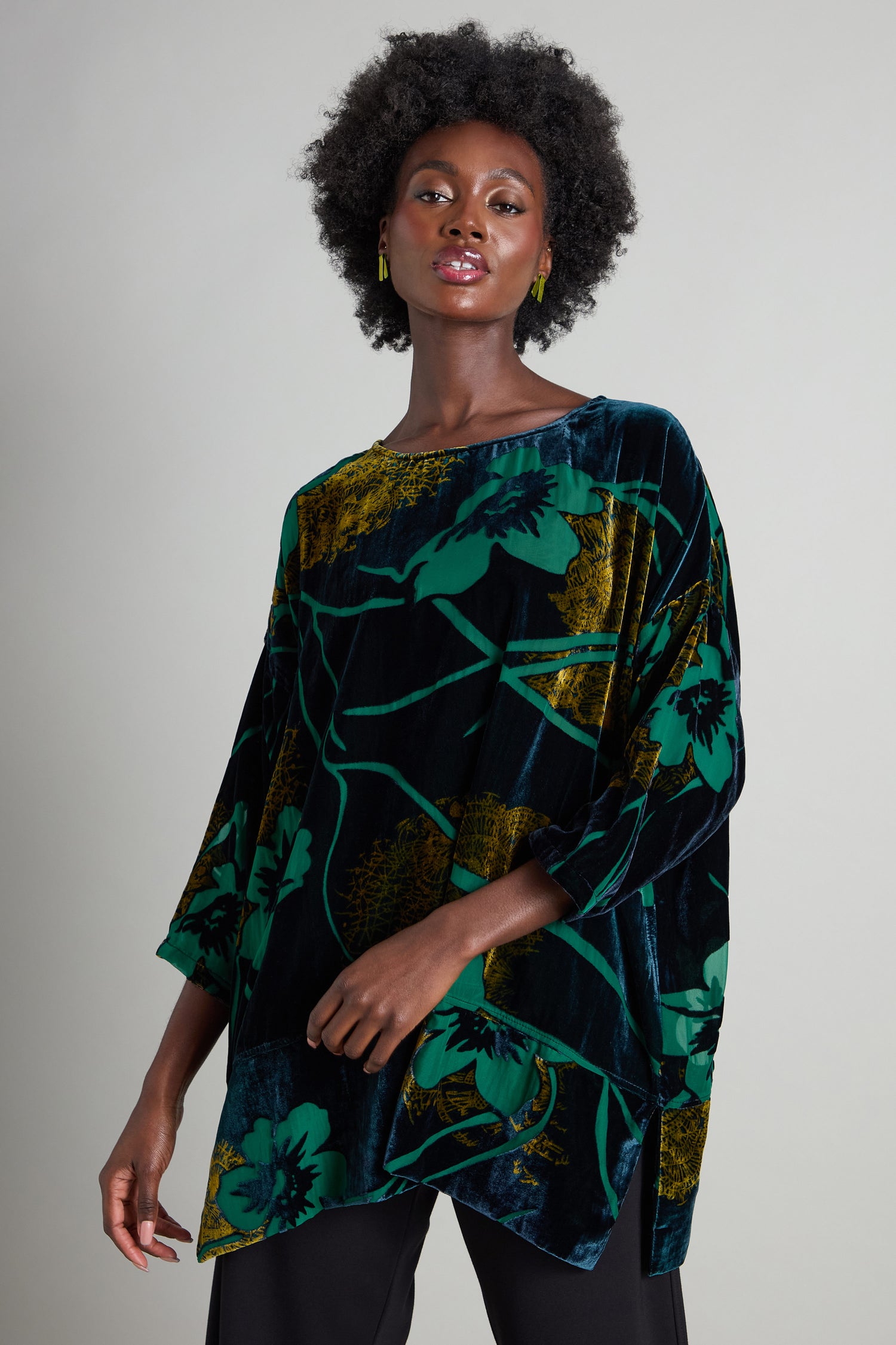 A person with curly hair confidently stands against a neutral background, wearing the Lily Devore Tunic embellished with green lily motifs and gold accents.