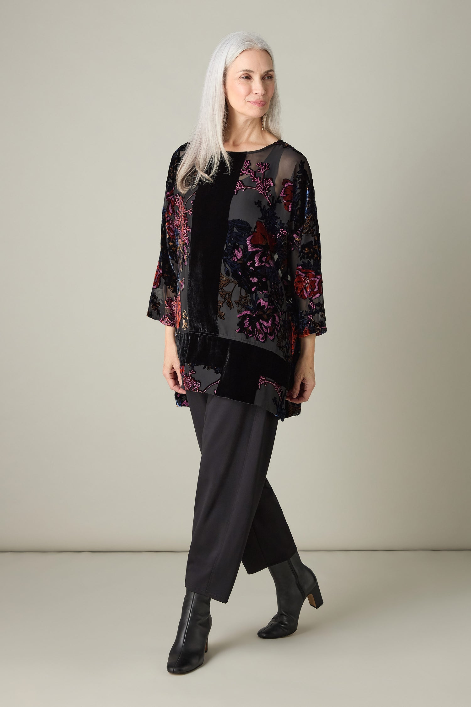 A person with long gray hair wearing a floral-patterned Cross Detail Devore Tunic along with black pants stands against a plain backdrop.