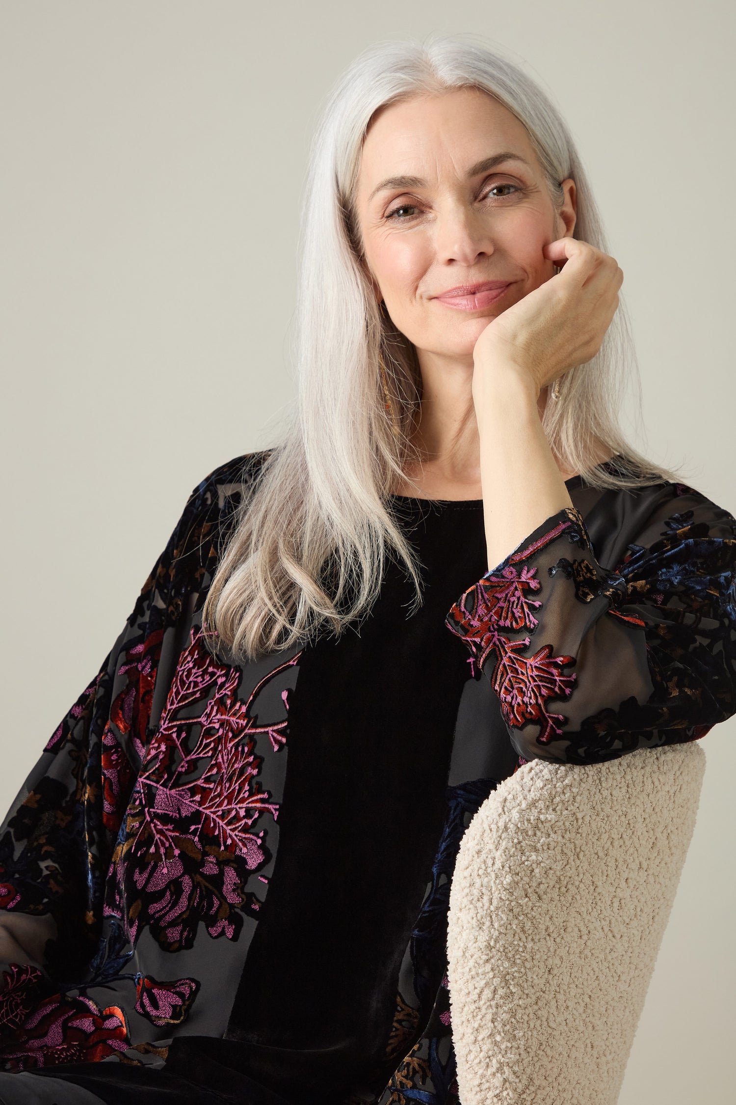 An older woman with long gray hair, wearing a Cross Detail Devore Tunic adorned with floral patterns, sits on a light-colored chair with her hand resting on her cheek.