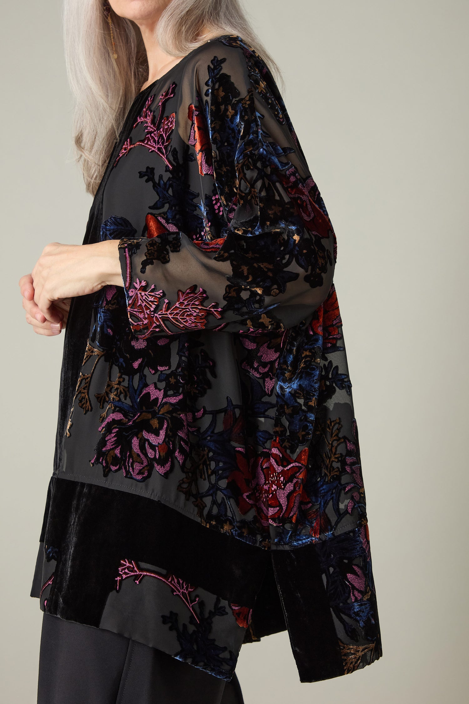 A person with long hair is wearing a Cross Detail Devore Tunic, which features a patterned, multicolored sheer blouse adorned with floral designs, and stands against a neutral background.