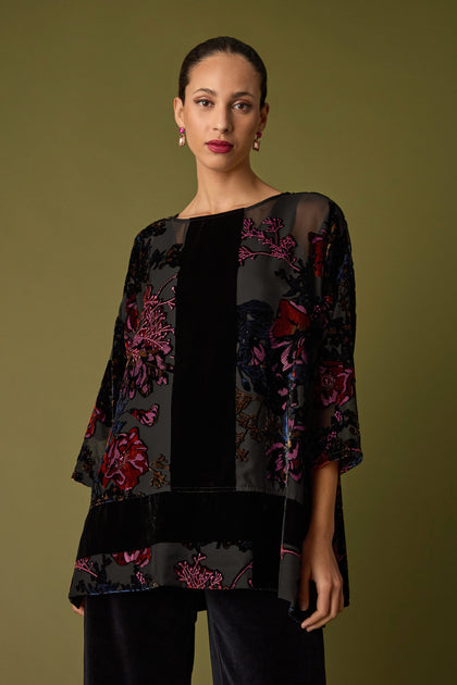 Dressed in a Cross Detail Devore Tunic featuring intricate floral patterns, paired with black pants, the person stands against a green backdrop. The velvet devoré fabric enhances the outfit's elegance.