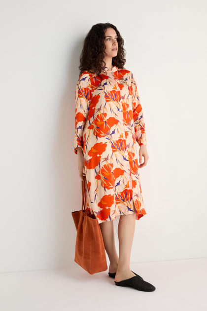 A woman stands against a white wall wearing the Japanese Floral Shirt Dress, featuring a knee-length, long-sleeve floral print with red, orange, and purple accents. She holds an orange tote bag and wears black flats.