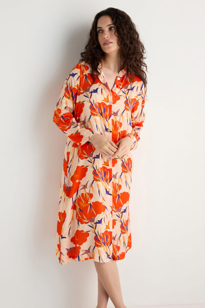 A woman stands against a white wall wearing the Japanese Floral Shirt Dress, a long-sleeve, button-down dress featuring a bold red and orange floral print.