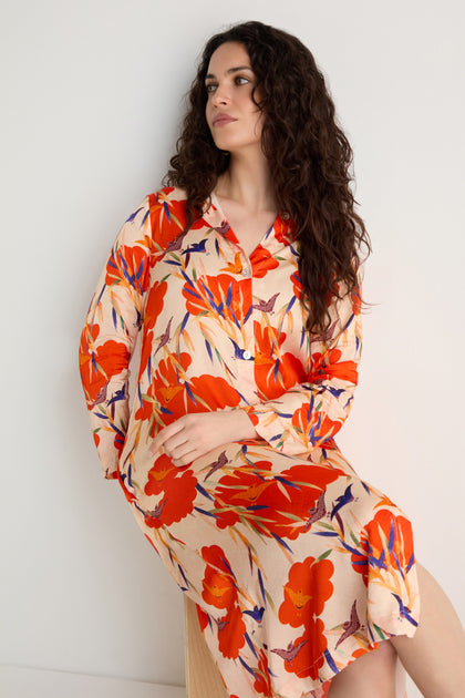 A woman with long curly hair wears the Japanese Floral Shirt Dress, a beige dress with bold red and orange blooms, as she sits against a white background.