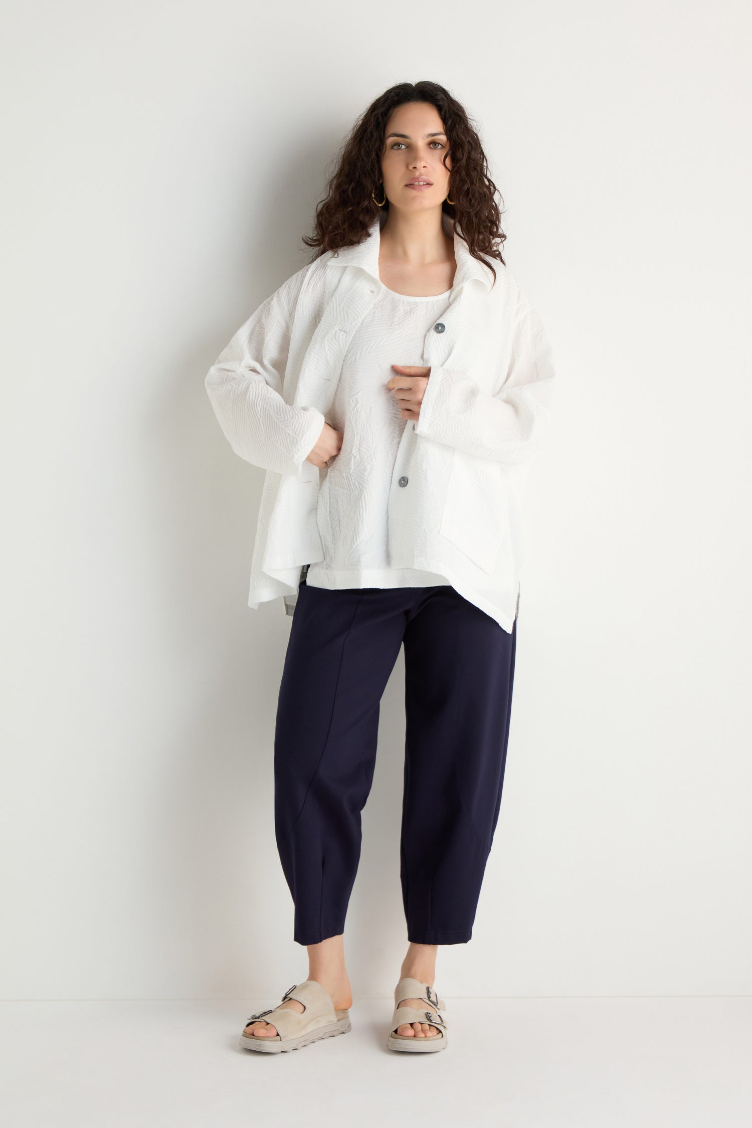 A woman stands against a white wall, wearing the Pleated Leaf Short Jacket over a white top, paired with navy blue pants and beige sandals.