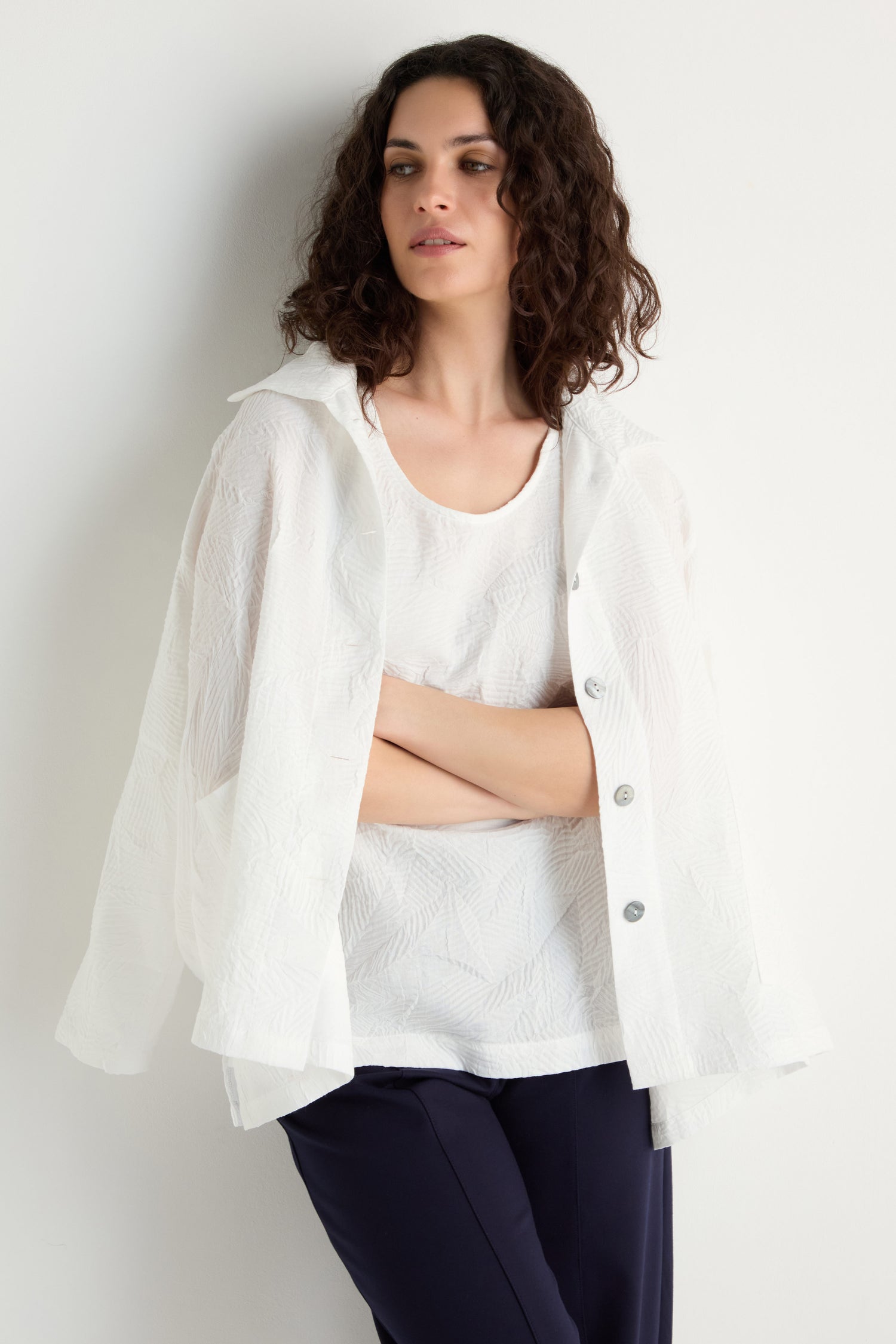 A woman with curly hair stands against a plain white wall, arms crossed, wearing the Pleated Leaf Short Jacket over a white top and dark pants.