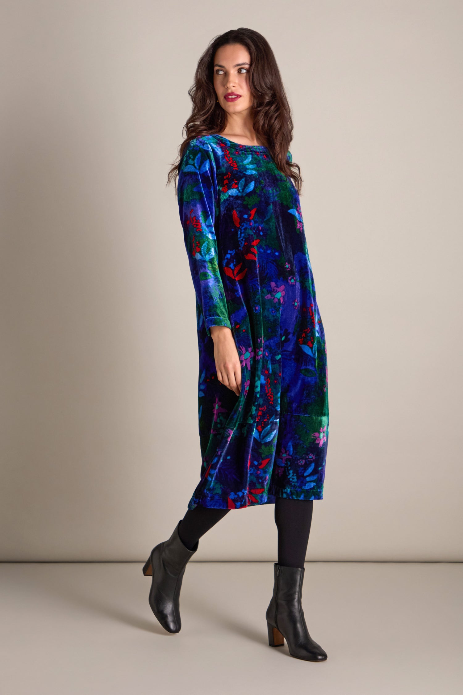 A woman wearing the Late Night Flora Velvet Bubble Dress, featuring a knee-length, long-sleeved blue and green floral print, complemented by black leggings and ankle boots, poses against a plain background, perfect for evening events.