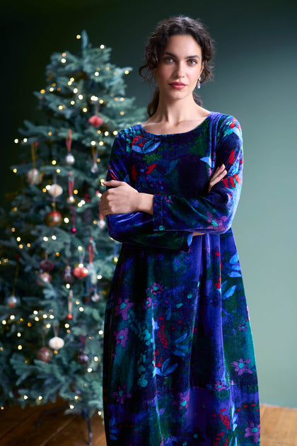 A person in a Late Night Flora Velvet Bubble Dress stands with arms crossed in front of a decorated Christmas tree adorned with lights and ornaments, perfectly ready for evening events.