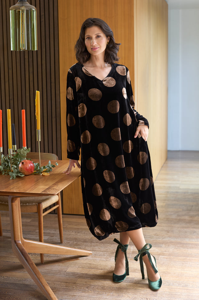 A woman stands indoors wearing the Silk Velvet Spot Bubble Dress, a long black dress with large brown polka dots, paired with green ribboned heels beside a wooden dining table set with candles and a pomegranate.