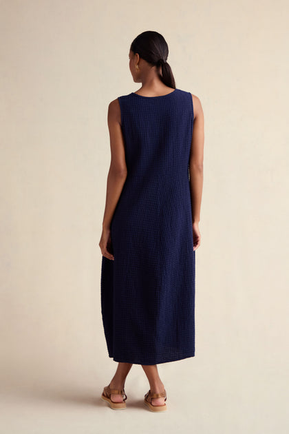 A person faces away from the camera, wearing the Mini Waffle Cotton Bubble Dress in navy blue with sandals, set against a plain light background.
