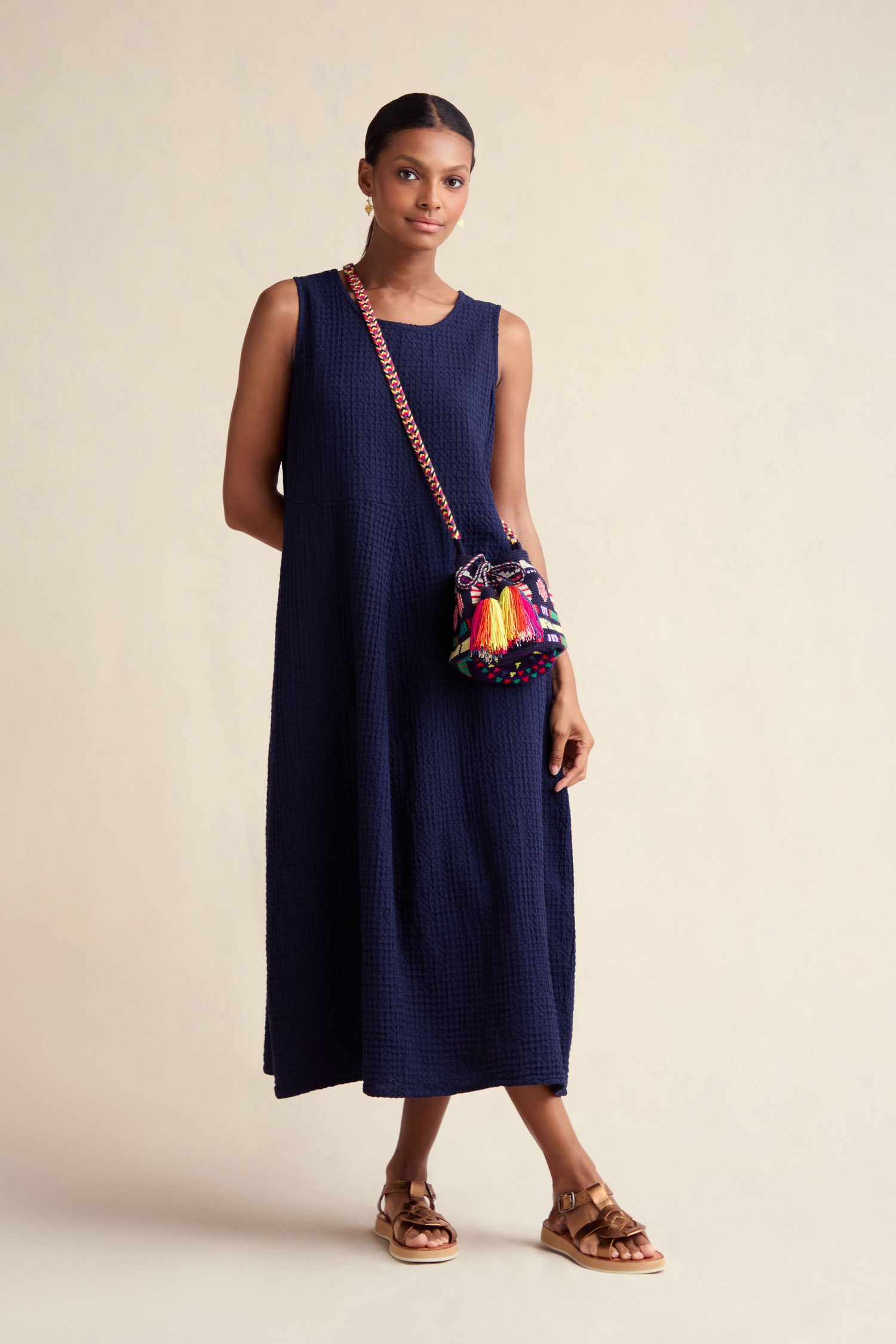 A woman wears the Mini Waffle Cotton Bubble Dress, a sleeveless navy blue midi, paired with brown sandals and a colorful crossbody bag against a plain light background.