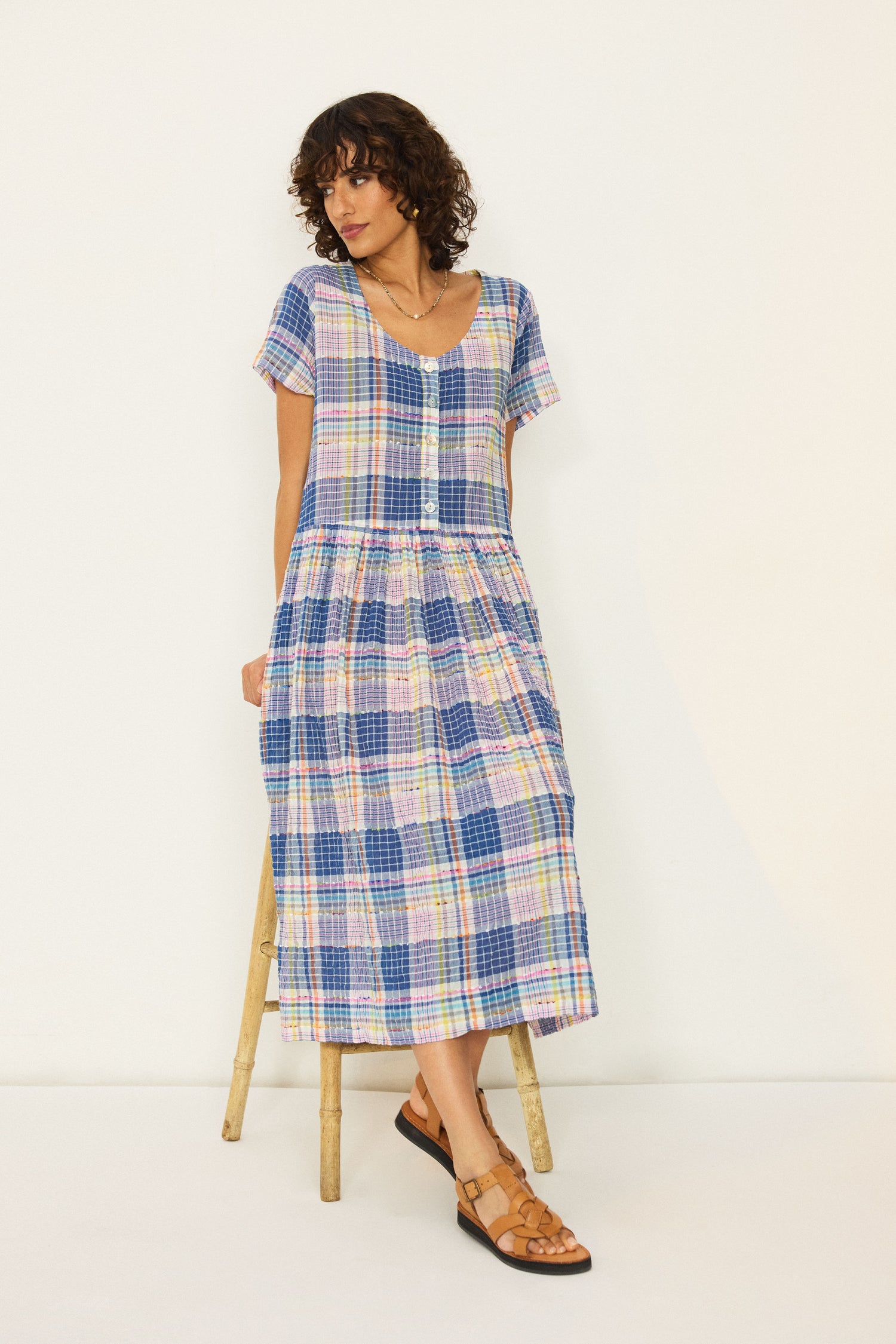 A woman stands against a plain background wearing the Crinkle Check Jacquard Dress—short-sleeved, v-neck, A-line with button details—paired with brown sandals.
