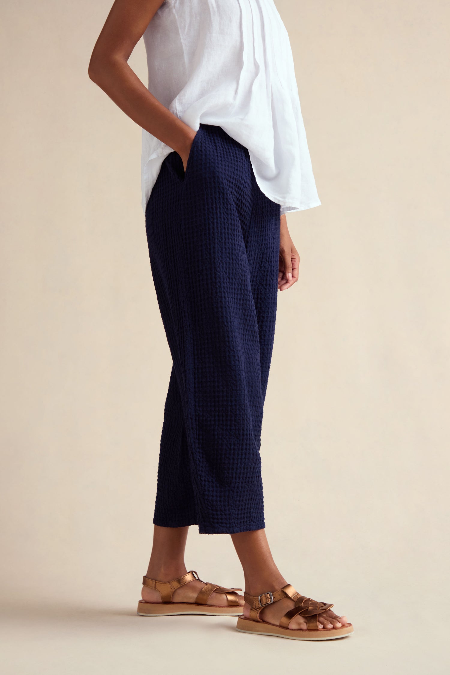 Wearing a white sleeveless top, tan sandals, and Mini Waffle Cotton Bubble Trouser in navy with a cropped length, a person stands with hands in pockets against a neutral background.