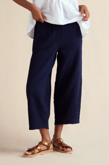 Person wearing Mini Waffle Cotton Bubble Trouser in navy, paired with a white top and tan sandals, standing against a neutral background. Only the lower half of the body is visible.