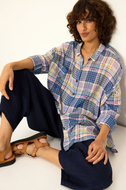 A person with curly brown hair sits against a white wall, wearing a loose plaid shirt, Mini Waffle Cotton Bubble Trousers in navy (cropped length), and tan sandals.