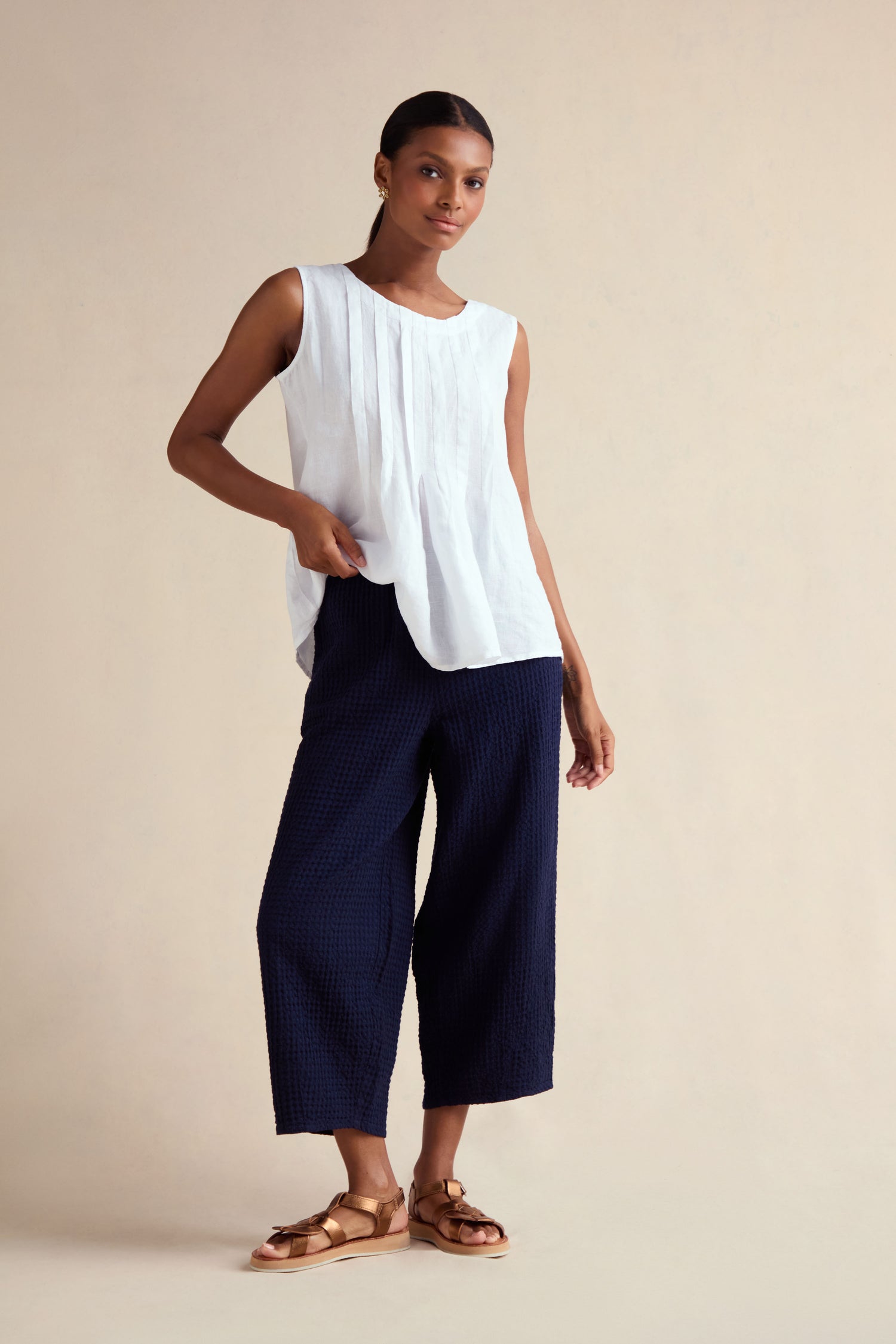 A woman stands against a beige background wearing a sleeveless white blouse, tan sandals, and Mini Waffle Cotton Bubble Trousers in cropped navy cotton.