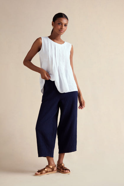 A woman stands against a beige background wearing a sleeveless white blouse, tan sandals, and Mini Waffle Cotton Bubble Trousers in cropped navy cotton.