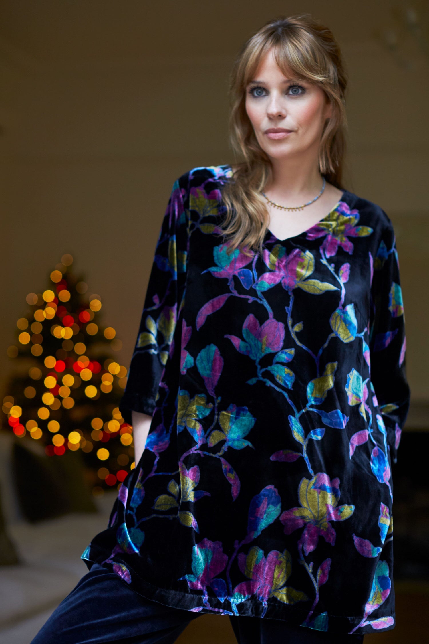 A woman, dressed in a Winter Magnolia Velvet Tunic, is posing gracefully in front of a Christmas tree adorned with Winter Magnolia floral decorations.