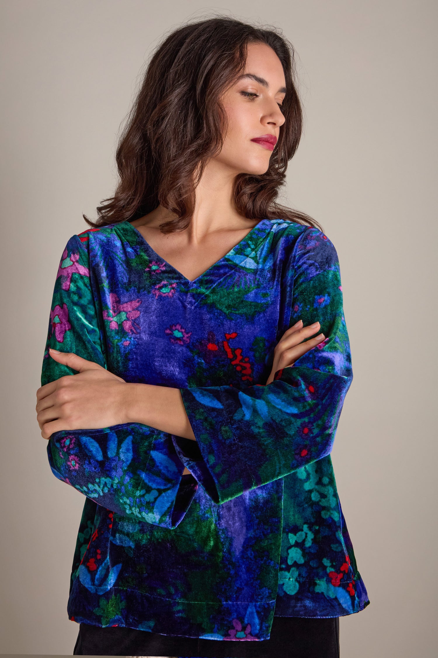 A person with long dark hair, clad in a Late Night Flora Velvet Top featuring a jewel-toned floral print, stands with arms crossed while glancing to the side.