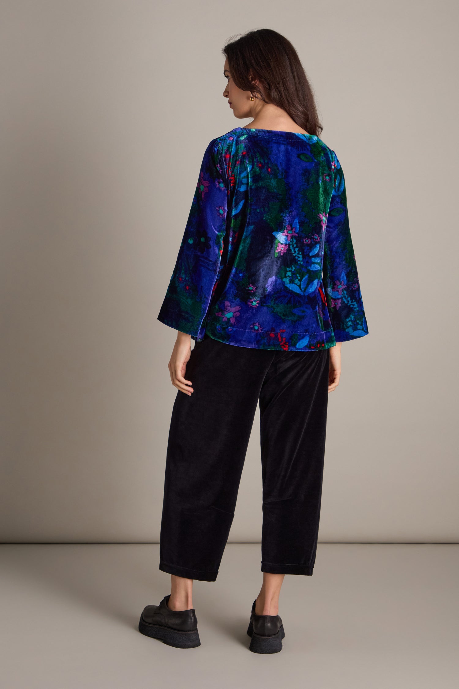 A woman is seen from behind, dressed in the eye-catching Late Night Flora Velvet Top featuring long sleeves, complemented by black velvet pants and shoes.