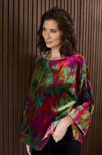 A woman with wavy dark hair stands before a wooden slat wall, wearing the Velvet Blurred Botanicals Top—a colorful, loose-fitting velvet piece featuring vibrant red, green, and yellow patterns.