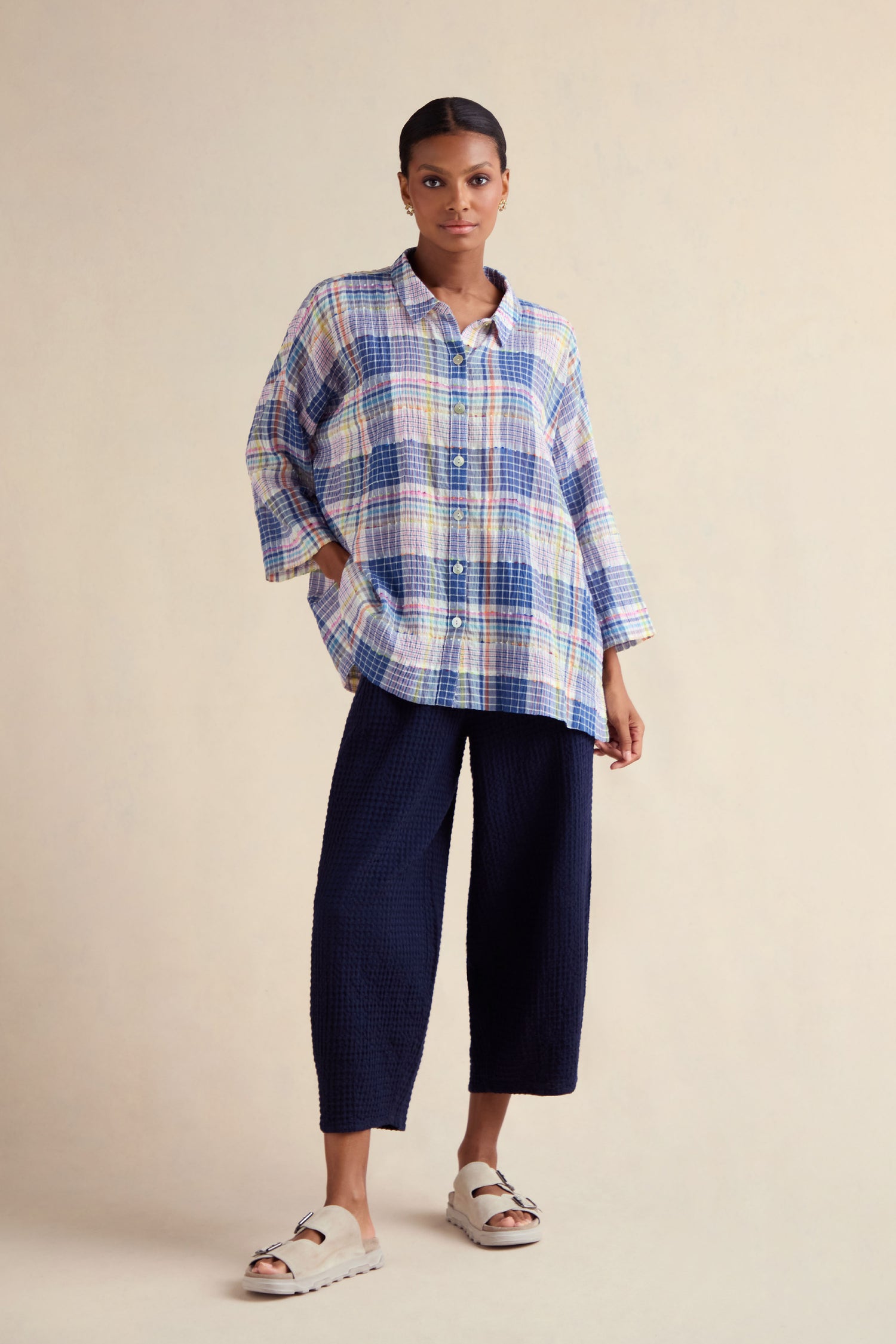 A woman stands against a plain background wearing a Crinkle Check Jacquard Shirt, textured navy wide-leg pants, and white sandals.