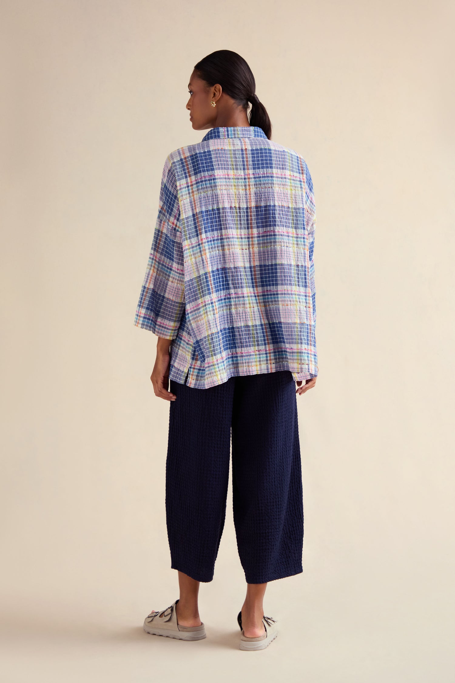 A woman stands with her back to the camera, wearing a Crinkle Check Jacquard Shirt in pastel shades, navy wide-leg trousers, and grey shoes against a plain beige background.